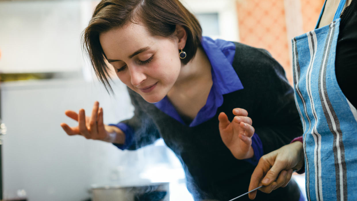  Woman leans over a steaming pan on a stovetop and wafts its scent towards her face. 