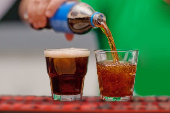 hand pours bottle of brown soda into two glasses.