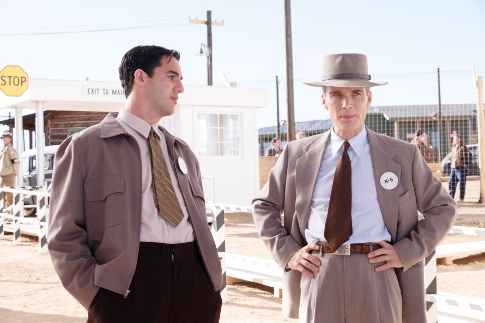Cillian Murphy as Oppenheimer stands next to Benny Safdie as Edward Teller in an outdoor setting with a "STOP" sign and buildings behind them