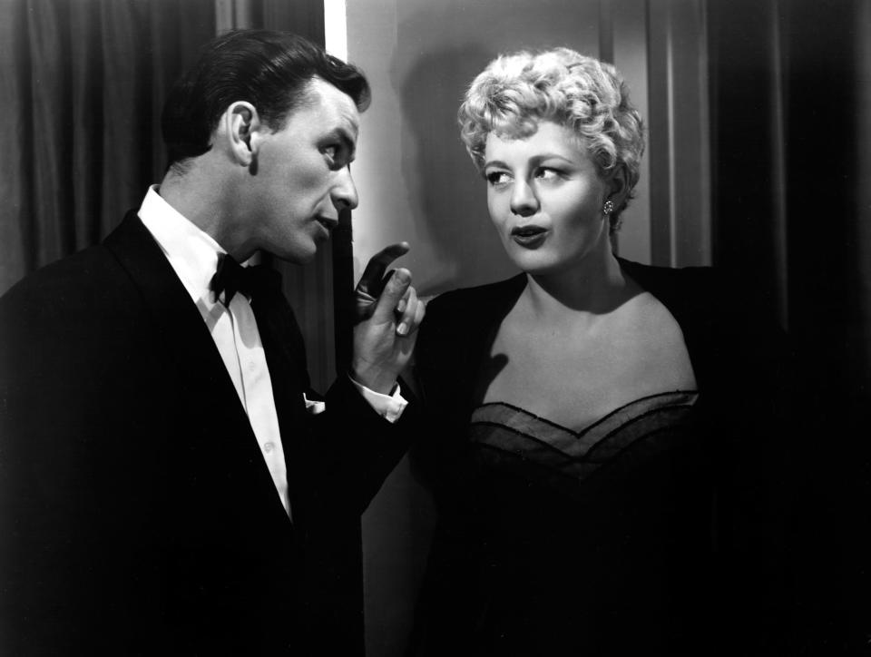 A man and a woman in formal attire are conversing in a hallway. The woman has wavy hair and is wearing a strapless dress with a blazer. The man is wearing a tuxedo