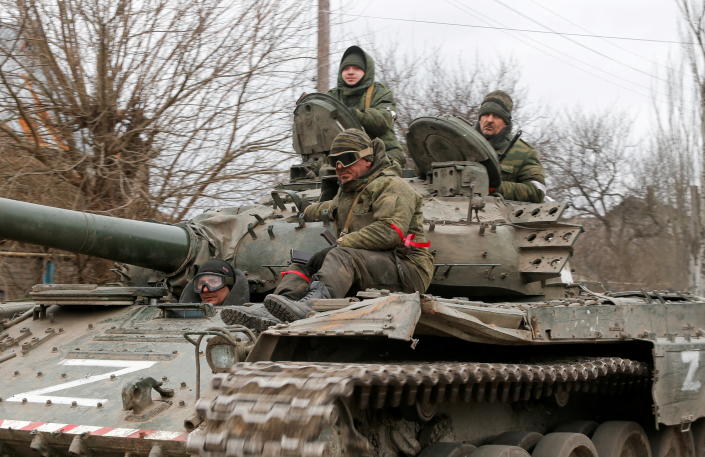 Three service members of pro-Russian troops in Ukraine in uniforms without insignia, one wearing red ribbons around his left arm and right leg, ride a tank marked 