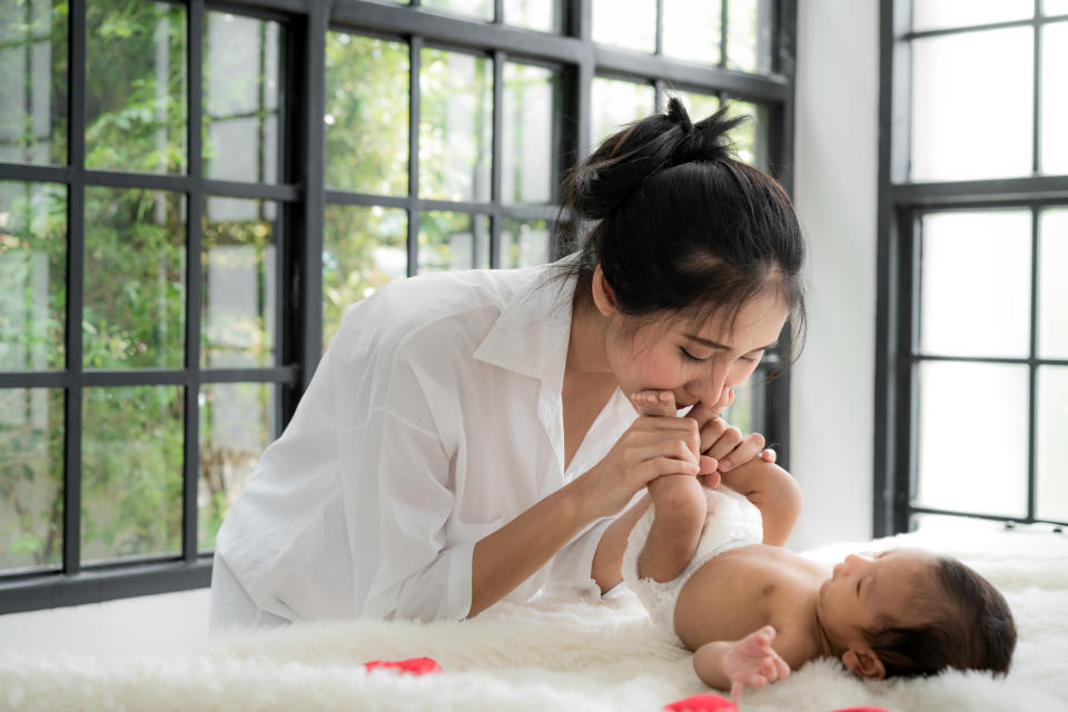 Asian happy young mother kissing her two months old daughter's feet in bed at home