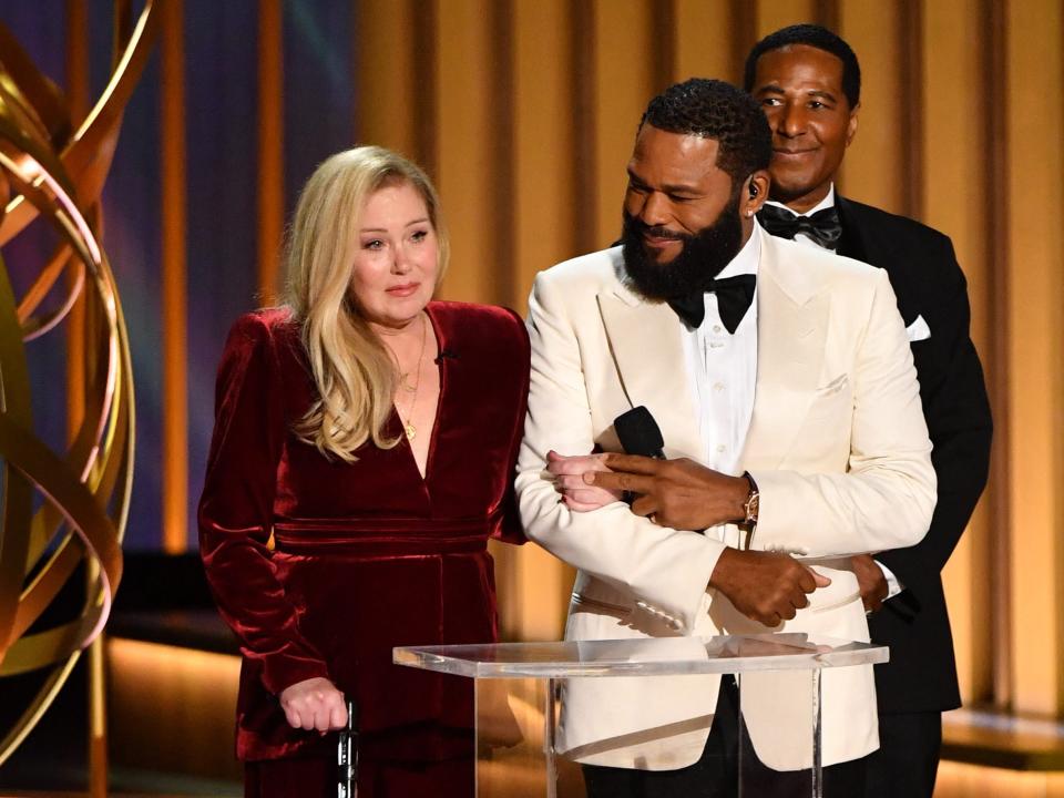 christina applegate in a red velvet dress with a black cane, on the arm of anthony anderson at a podium on the emmys stage