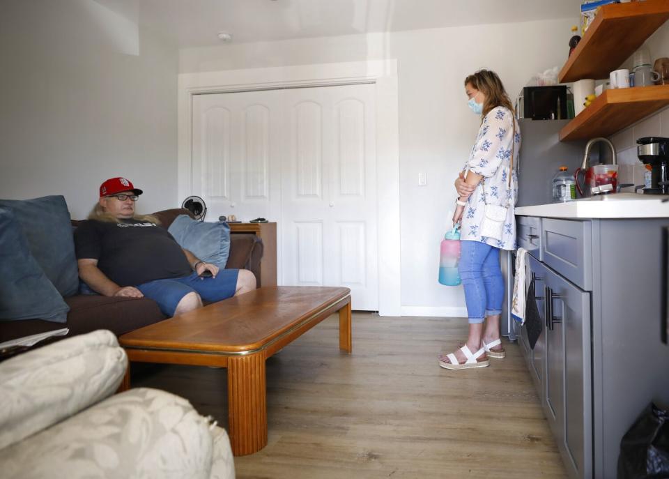 A woman stands talking to a man sitting on a couch.