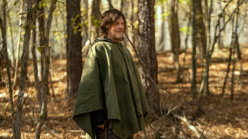 Norman Reedus as Daryl Dixon on the walking dead stands in a between trees with brown leaves on the ground while wearing a poncho