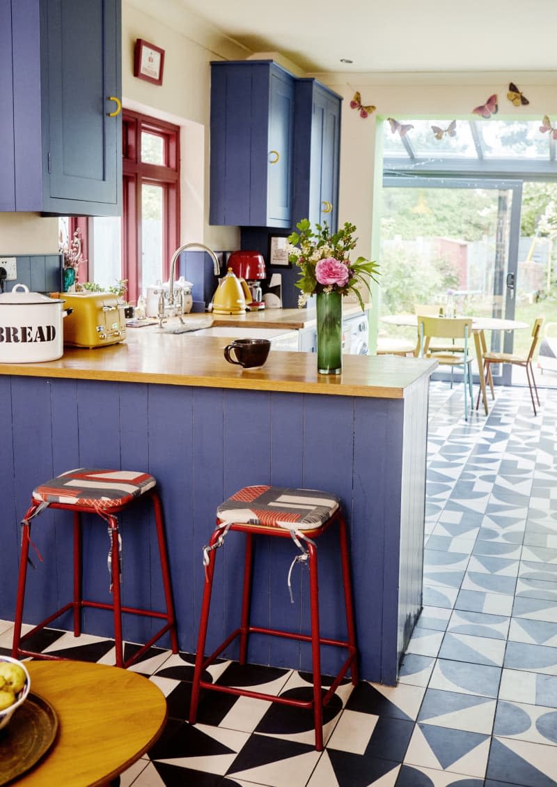 open door way, blue cabinets, butcher block countertop, white and black geometric tile floor, oven, wood cutting board, butterfly garland, flowers, yellow electric tea kettle, yellow, toaster, red coffee maker, greenhouse,  sliding door, glass ceiling, red metal bar stools, yellow chairs, oval table, bread jar, red window frames