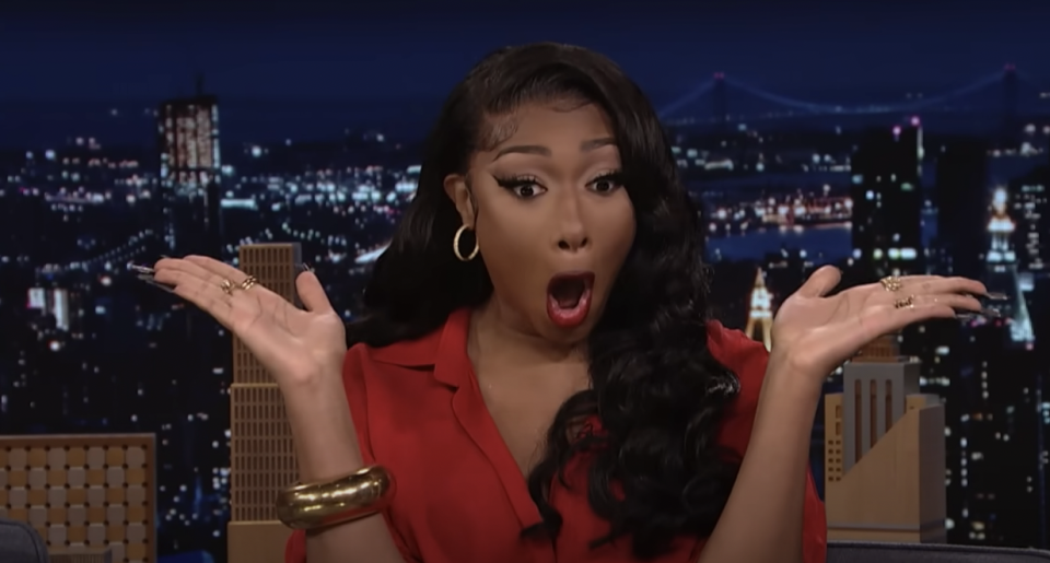 Megan Thee Stallion on a talk show set, wearing a red blouse, gold hoop earrings, and bracelets, with a surprised expression and hands raised