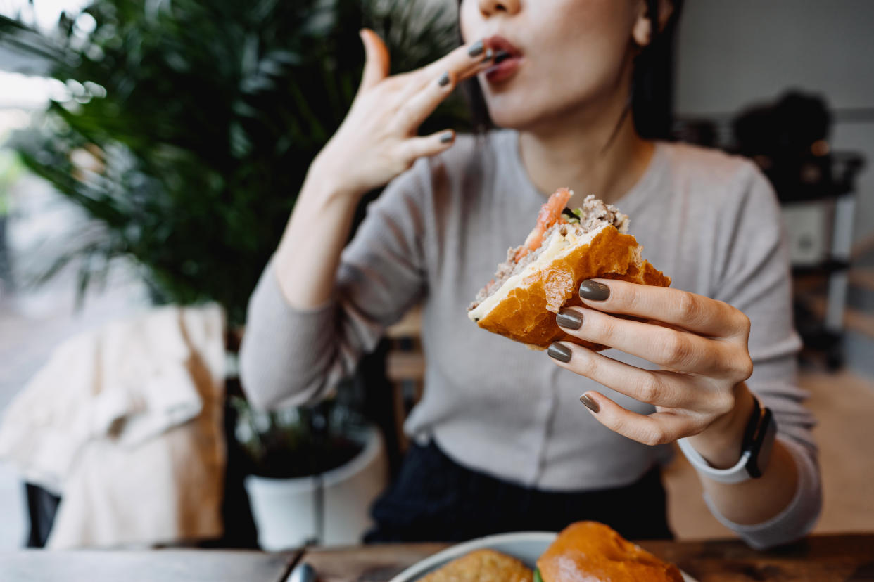Cropped shot, mid-section of young Asian woman eating freshly made delicious cheeseburger in a cafe, licking her fingers. Enjoying her lunch! People and food concept