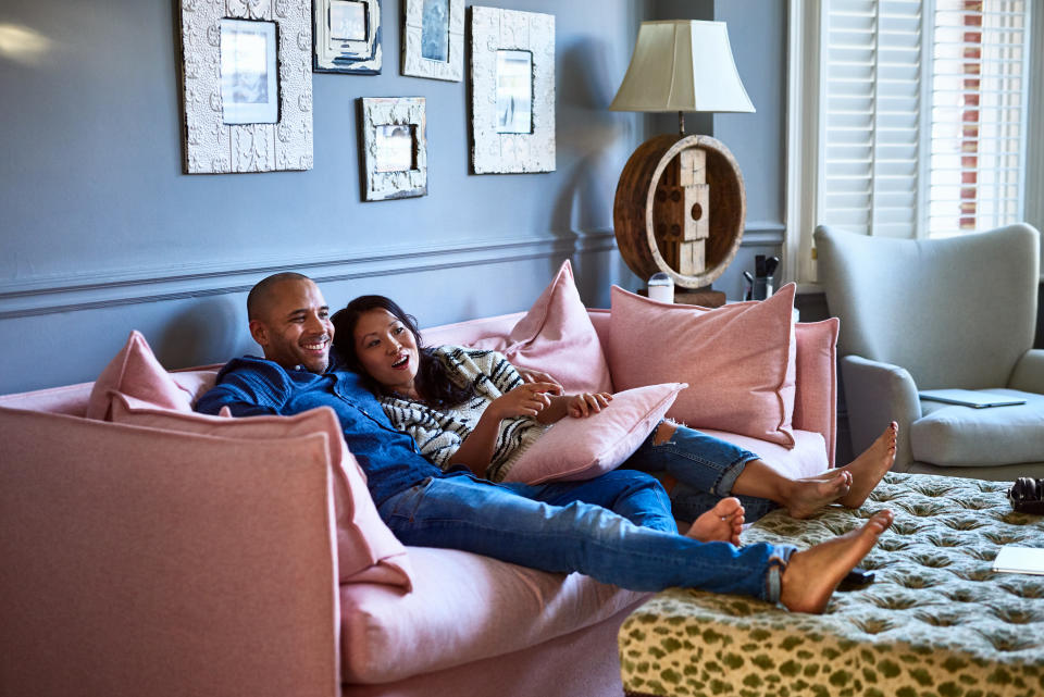 Mature man with arm around Vietnamese woman, sitting with feet up and smiling, woman pointing to TV, entertainment, relaxation, togetherness