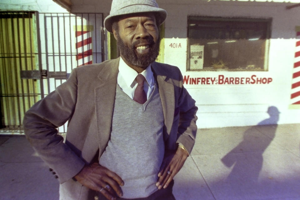 oprah winfrey's dad veron in a gray hat, burgundy tie, grey shirt and brown jacket in front of his barbershop 