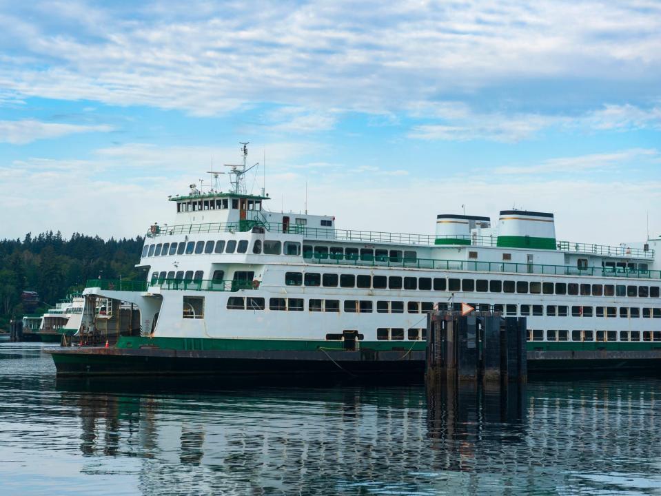 A Washington State Ferry boat at the Bainbridge Island Terminal. The boat is mostly white with green detailing and sits in water with pine trees and blue skies in the background