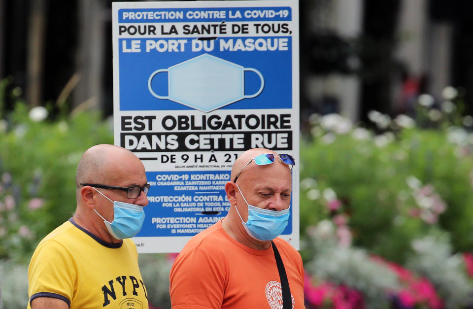 People wearing face masks to protect against coronavirus walk past a sign stating face coverings are required in the street, in Bayonne, southwestern France, Wednesday, July 28, 2021. Local authorities in France are re-imposing mask mandates and other virus restrictions because of fast-growing infections with the delta variant, which is causing COVID-19 hospitalizations in France to rise again. (AP Photo/Bob Edme)