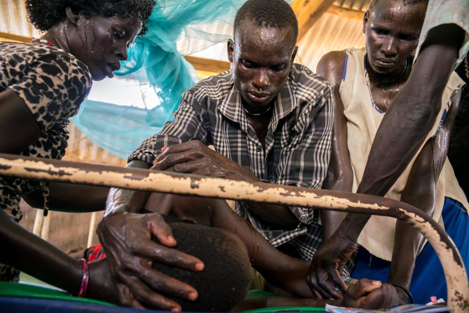 A semi-conscious man is cared for by several adults who hover over him.