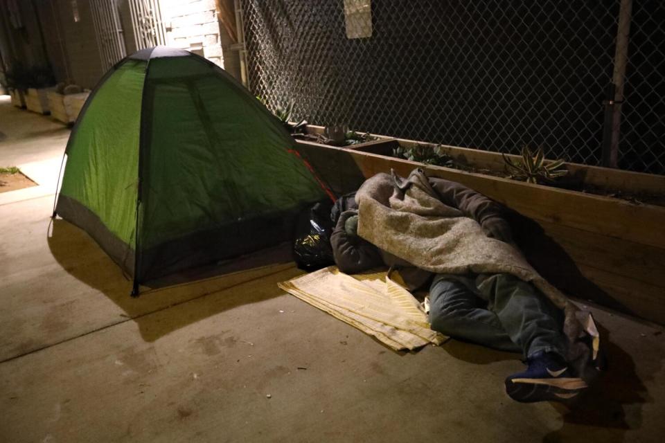 A homeless man sleeps on a sidewalk next to a small green tent