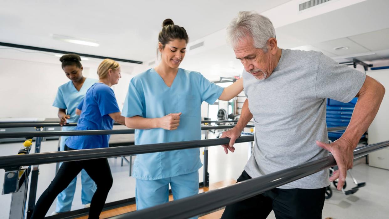 Diverse team of physiotherapist helping senior patients walk between parallel bars all looking happy and smiling.