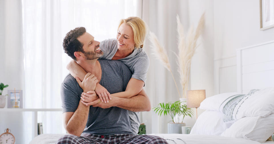 A happy couple embraces on a bed, smiling at each other lovingly. Both are dressed in casual, comfortable clothing