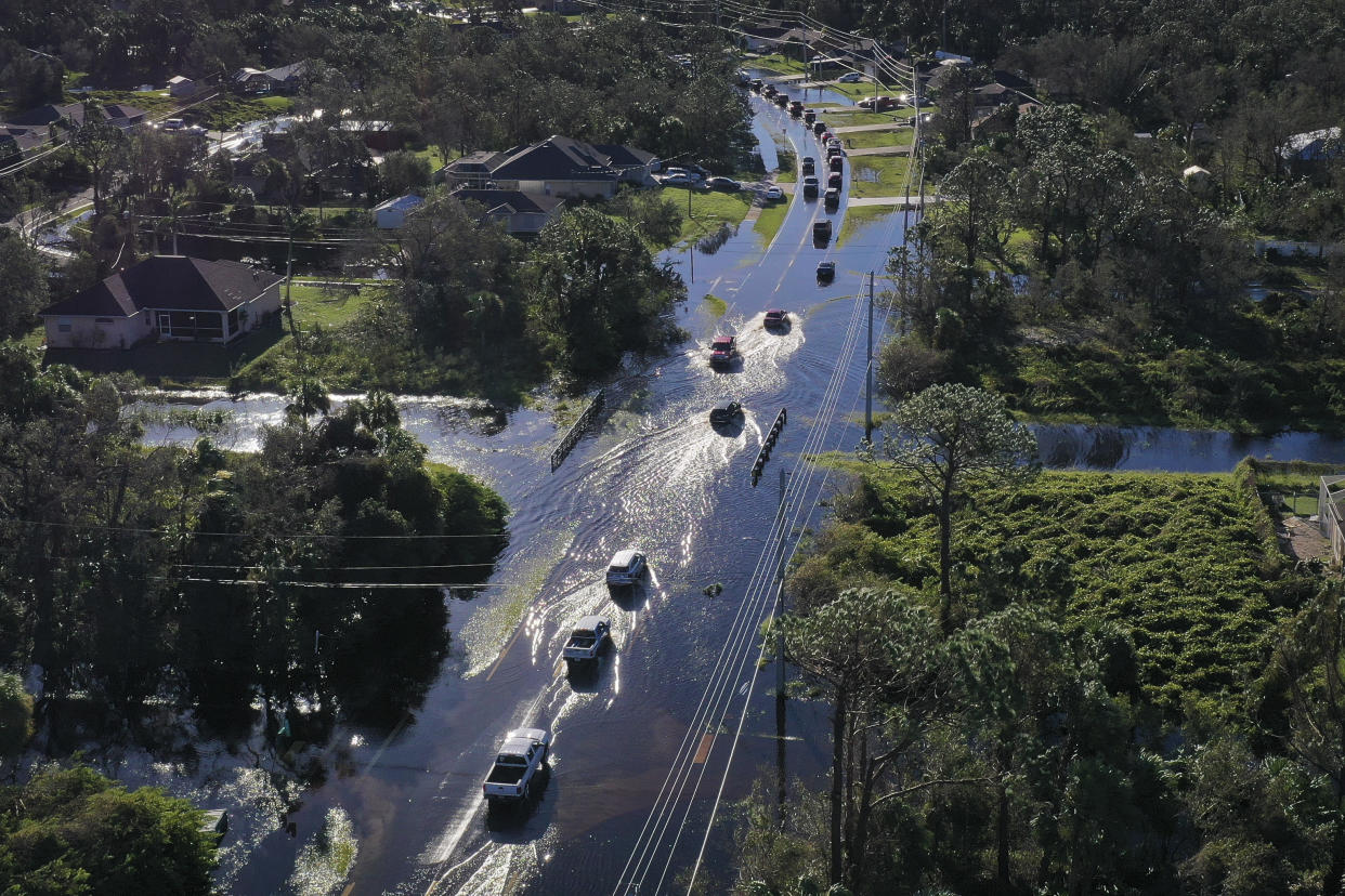 An aerial view of a line of vehicles leaving a wake as they drive through several inches of water on a flooded roadway.