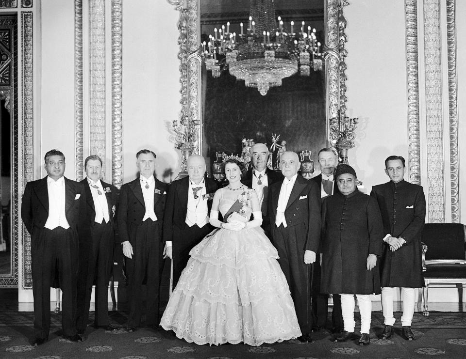 The officials in formal dress line up around the queen, who is wearing a crinoline and tiara, under a large chandelier.