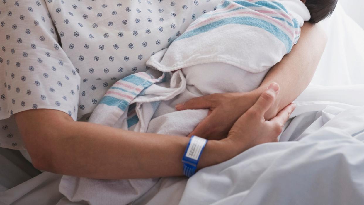  close up photo of a mother's arms holding a baby; the woman's skin is  tan and she's wearing a hospital bracelet on her wrist and a hospital gown 