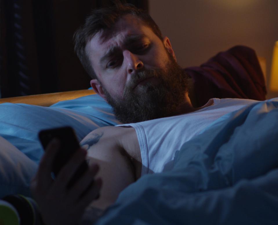 A man with a beard is lying in bed at night, looking at his phone. He appears to be tired or concerned. A bedside lamp is on beside him