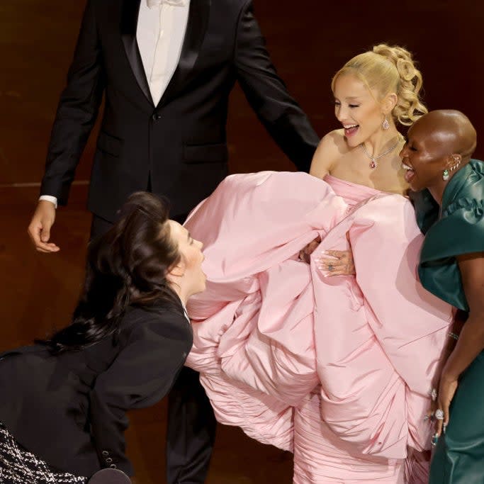 Four people laughing and interacting at an event; one in a voluminous pink gown, another in a green ensemble