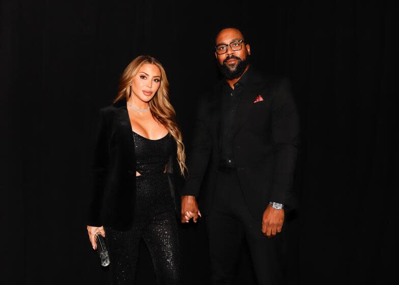 A woman with light brown hair in a black outfit holding hands with a Black man in glasses and a black outfit