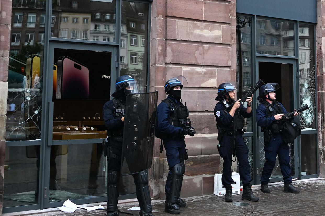 French police officers in riot gear stand guard and hold tear gas canister launchers next to the facade of a damaged Apple Store at Place Kleber, in Strasbourg, eastern France, on 30 June 2023 (AFP via Getty Images)