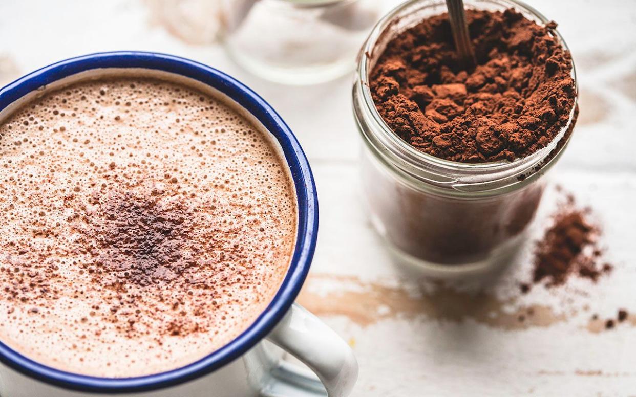 A warm cup of Hot Chocolate, on a rustic white-washed timber table top, with a jar of cacao on the side.