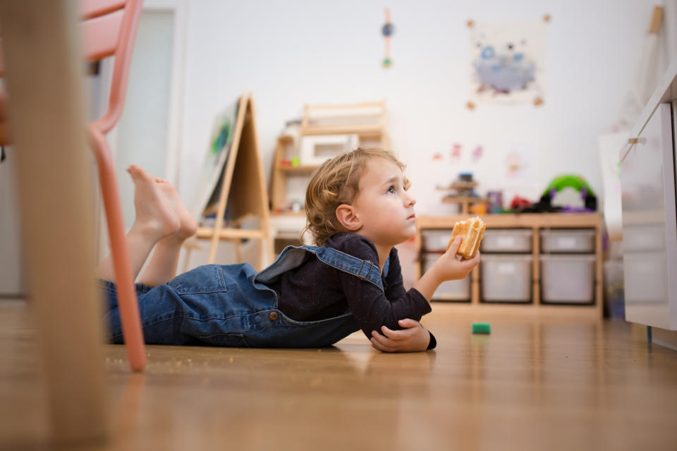 A young child in overalls lays on the floor, intently watching something while holding a sandwich. Shelving and toys are visible in the background