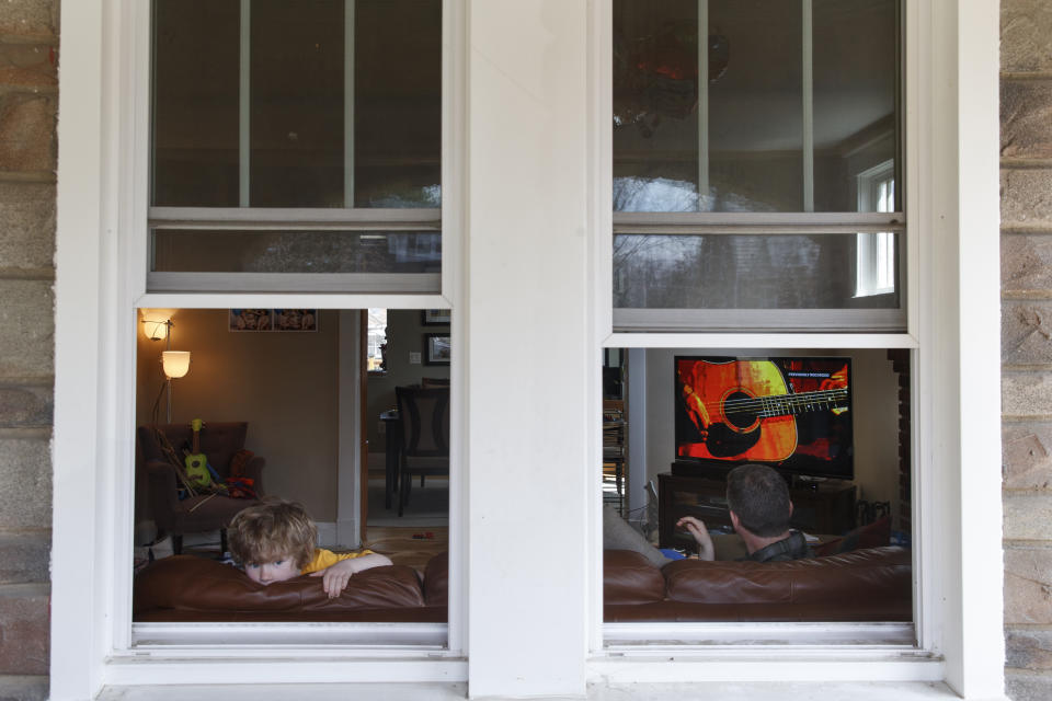 Ethan McIver, 4, son of U.S. Army Field Band member Sgt. Major Robert McIver, Jr., right, looks out the window of their Catonsville, Md., home, Thursday, March 26, 2020. The family is staying home because of the coronavirus outbreak. Inside on the television the U.S. Army Field Band's daily "We Stand Ready" virtual concert series from Fort George G. Meade is playing. The Army Field Band's mission is to bring the military's story to the American people. And they're not letting the coronavirus get in the way. (AP Photo/Carolyn Kaster)