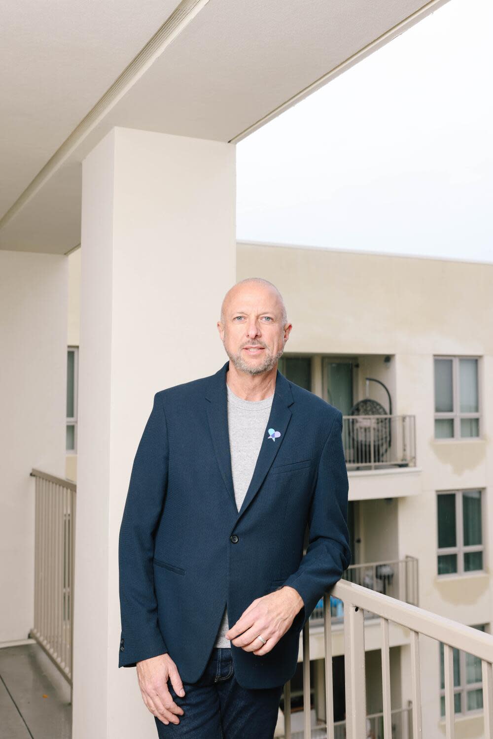 A man in a blue suit stands in an outdoor corridor in the middle of a beige apartment complex.