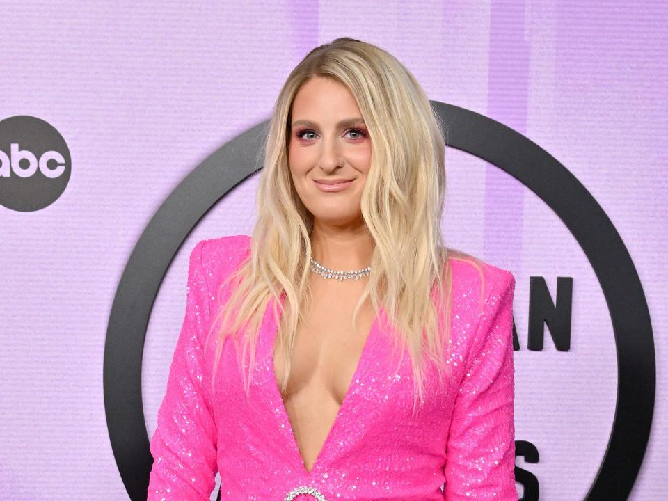 meghan trainor wearing a deep v, bright pink suit that exposes the inner curve of her breasts. her hair is lightly wavy, and she's standing on the american music awards red carpet