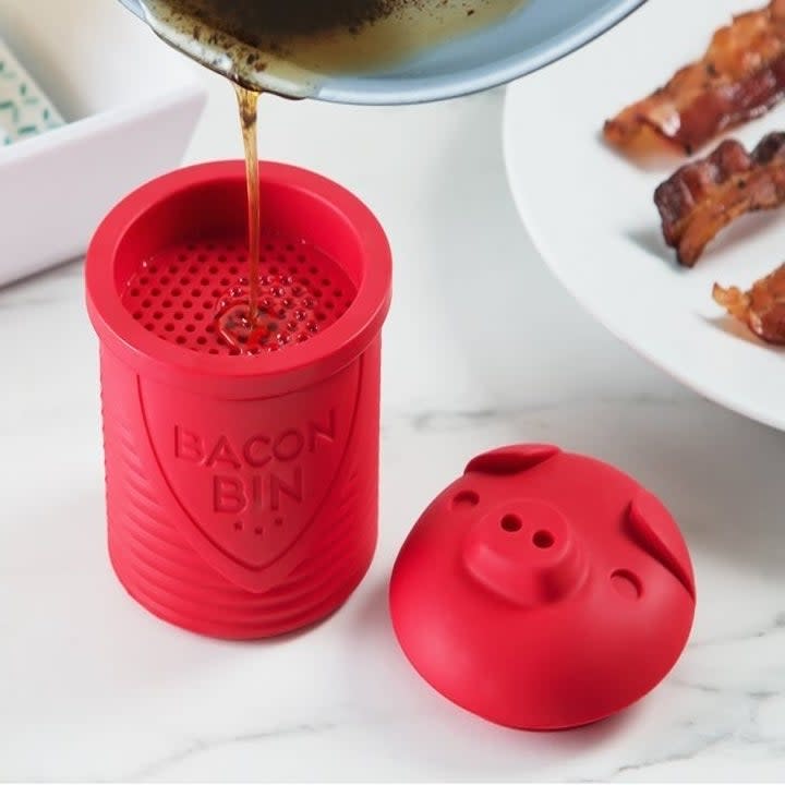 Oil being poured strained into the red silicon bacon pin sitting next to the matching silicone lid shaped like a pig's head 