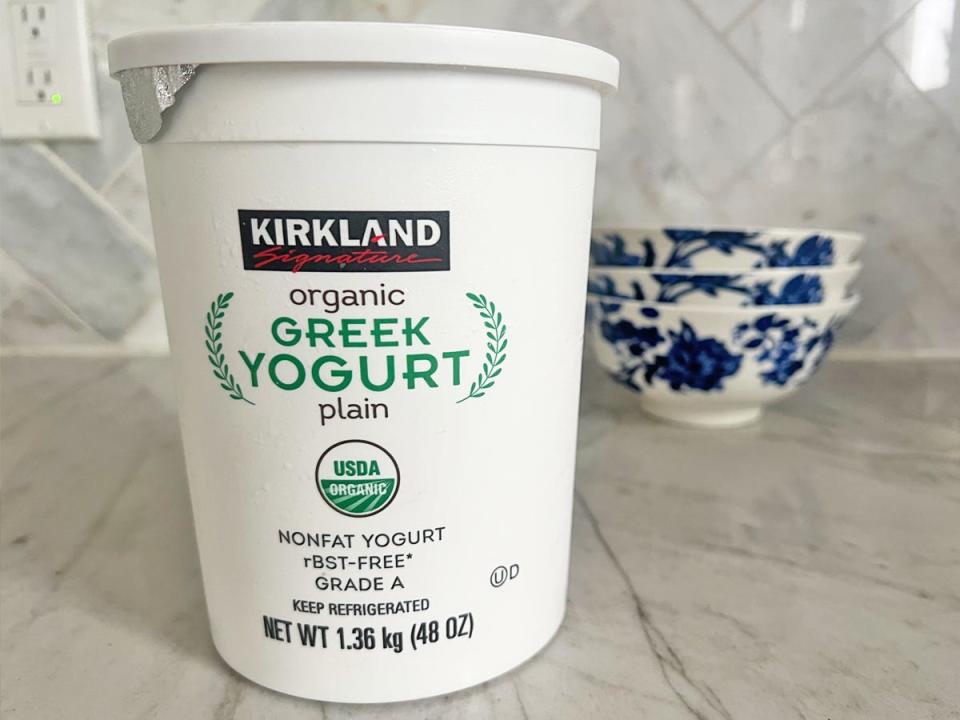 A large white container with Kirkland Signature and black and green text spelling out "organic Greek yogurt plain" on a gray countertop with blue-and-white bowls in the background