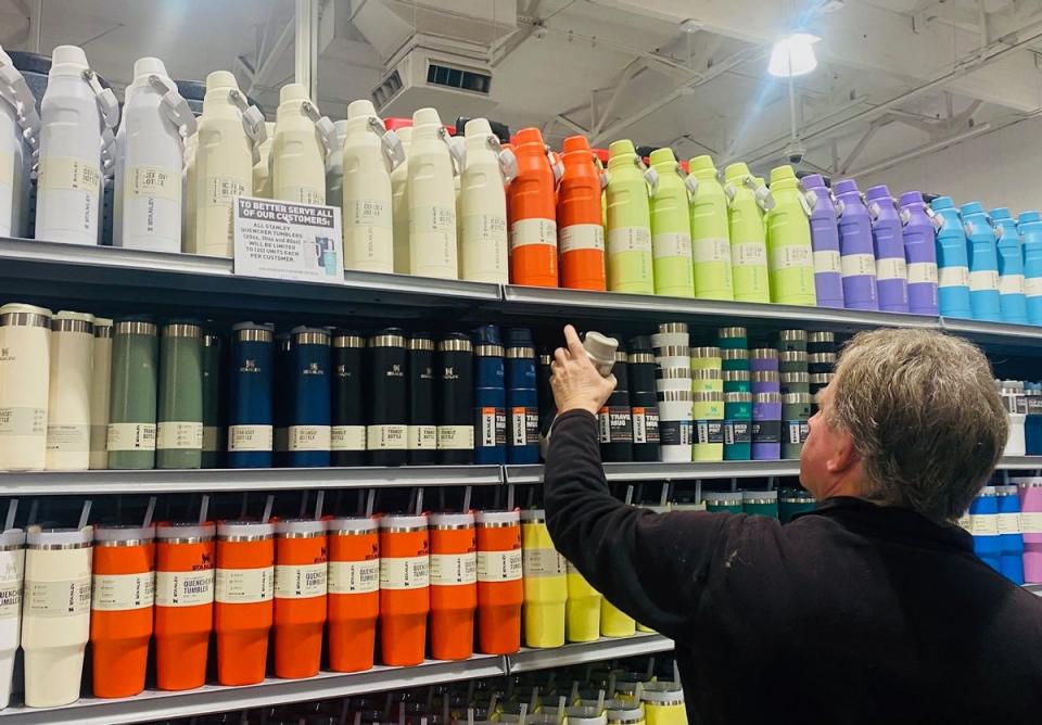 A white man stands before water tumblers in a range of different colors. A sign limits the amount that patrons can buy at the store. 