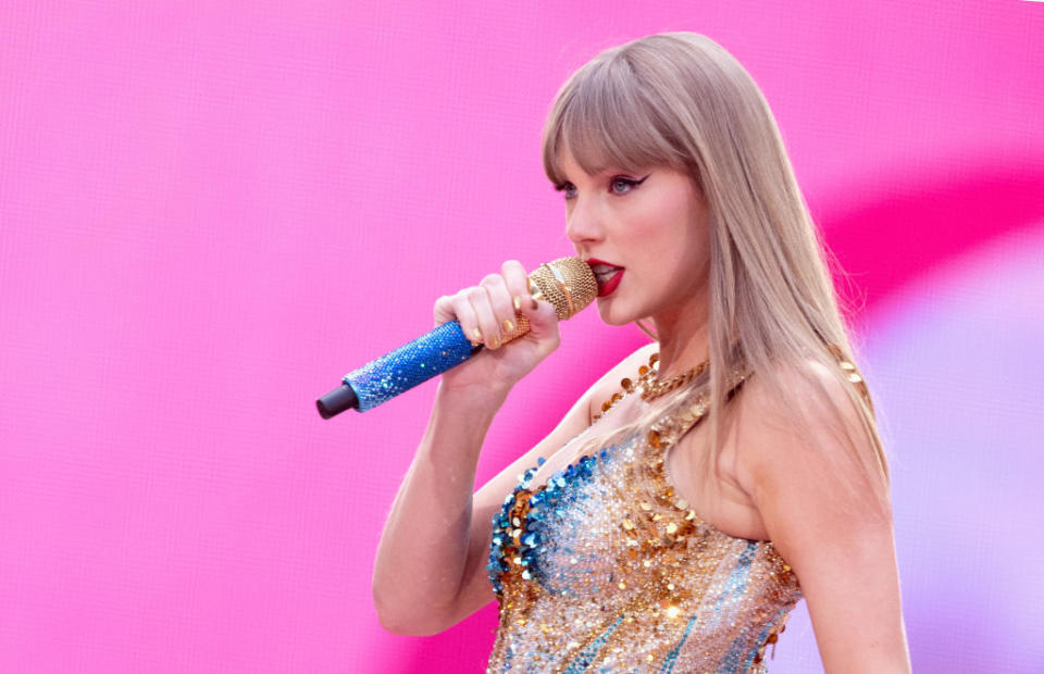 Taylor Swift performing on stage, holding a sparkling blue and gold microphone, wearing a sequined outfit with a gradient pattern
