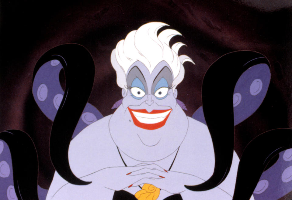 Ursula from "The Little Mermaid" smiling mischievously with her tentacles visible in the background. Her hands are folded, and she has white hair and bright red lipstick