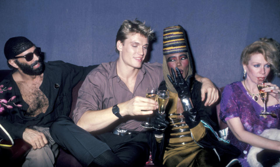 Group of four stylish individuals seated, holding drinks, in a social setting with vibrant fashion including sequins and unique headwear