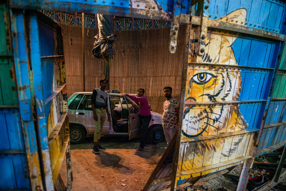 Three men stand in the "Well of Death" by a silver car with the doors open. The photography is taking the picture from outside of the structure through the door to the arena. There is a tiger painted on the outside wall.