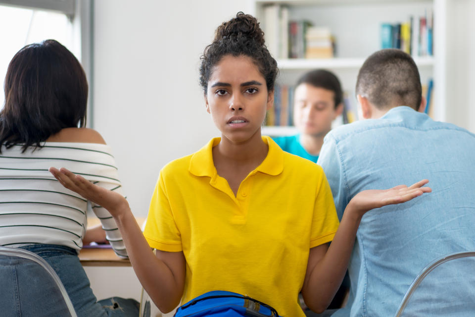 A young student in a yellow shirt holds out both her hands, palm up, as if to say: What does this mean?