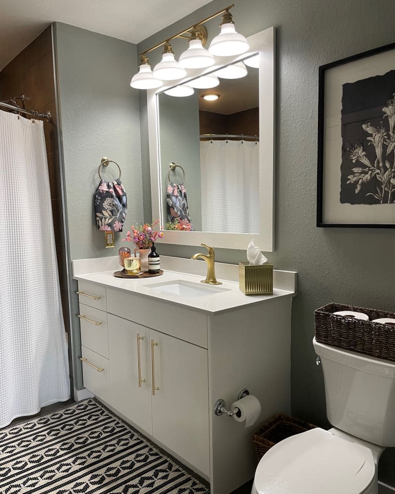 Wood tray holding accessories and gold tissue box atop white sink with gold faucet and white cabinetry under white framed mirror in sage bathroom with floral towel and art and graphic rug.