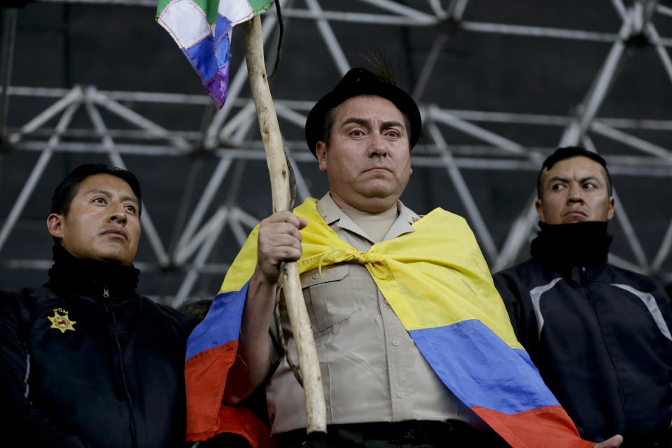 Col. Cristian Rueda Ramos, one of several police officers who has been detained by anti-governments protesters, is made to hold an indigenous banner, don a national flag as a cape and a fedora-styled hat, while presented on a stage at the Casa de Cultura in Quito, Ecuador, Thursday, Oct. 10, 2019. An indigenous leader and four other people have died in unrest in Ecuador since last week, the public defender's office said Thursday. (AP Photo/Fernando Vergara)