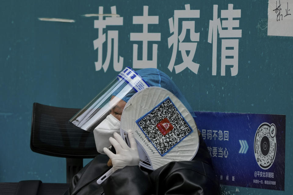 FILE - Holding a fan with the health check QR code printed on it, a worker in a protective suit talks on her phone at a coronavirus testing site in Beijing, Wednesday, Nov. 9, 2022. The sign in background reads: "Fight the epidemic." (AP Photo/Andy Wong, File)