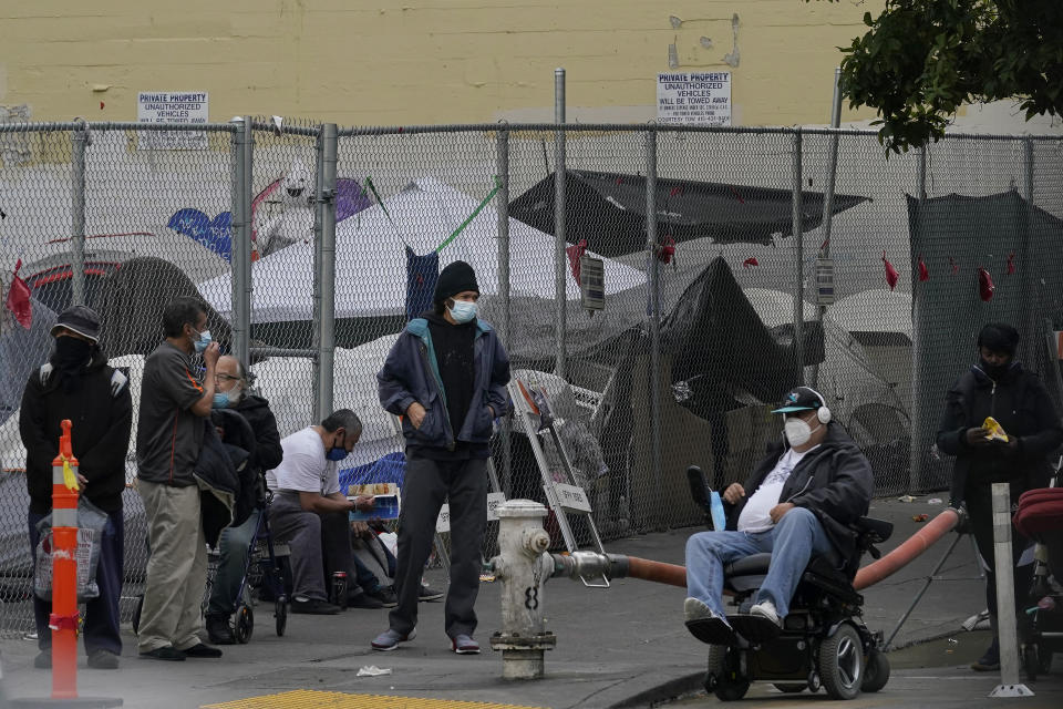 People linger on a street corner in front of tents set up in a fenced lot in San Francisco, Saturday, Nov. 21, 2020. Some counties in California are pushing ahead with plans to wind down a program that's housed homeless people in hotel rooms amid the pandemic, despite an emergency cash infusion from the state aimed at preventing those same residents from returning to the streets in cold, rainy weather as the virus surges. (AP Photo/Jeff Chiu)