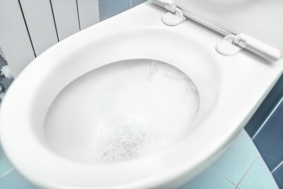 An open white porcelain toilet on teal tile being flushed.