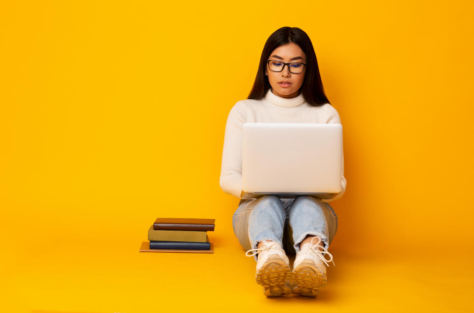 Asian Student Girl Using Laptop Studying Preparing For Tests And Exams Sitting With Books On Floor Over Yellow Studio Background.