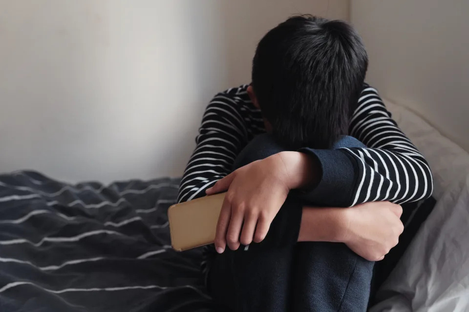 A sad child sitting on his bed with his head down and a phone in his hand