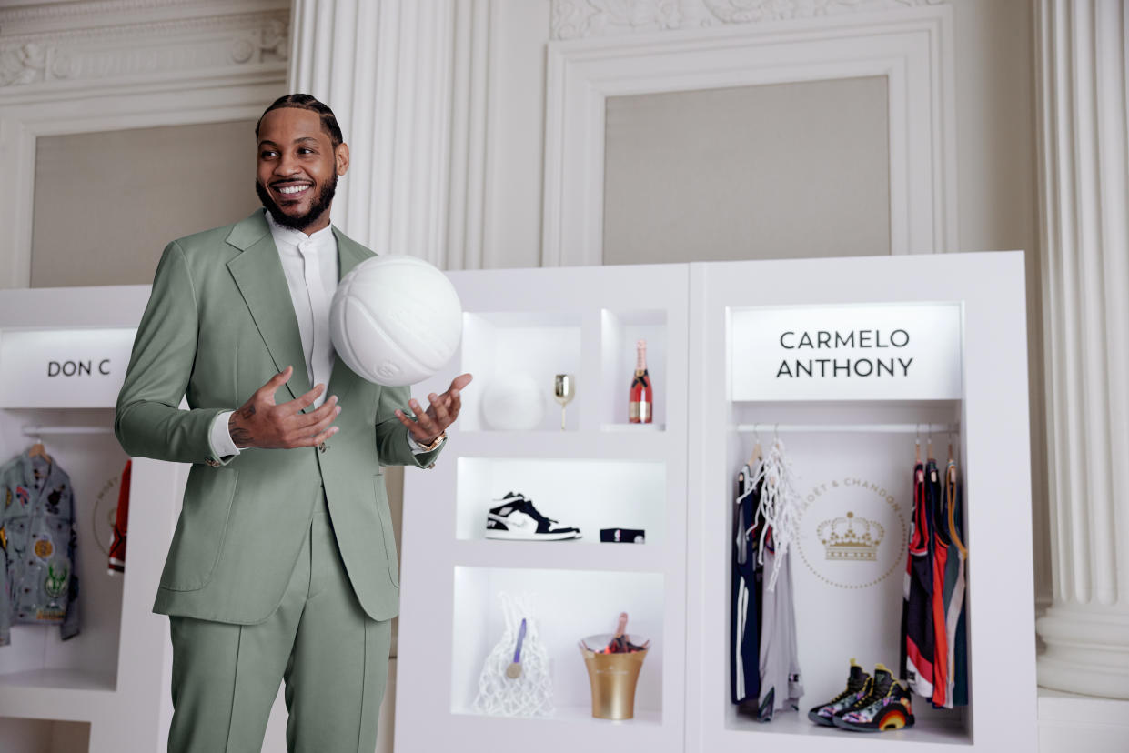Carmelo Anthony holding a basketball smiling n front of a locker room of shoes, champagne and jerseys.