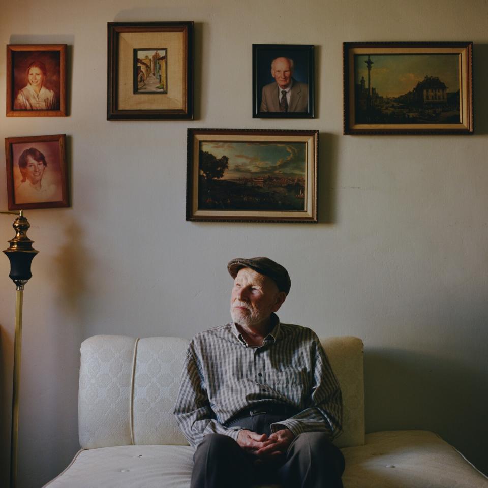 A man sits on a couch under pictures and paintings.