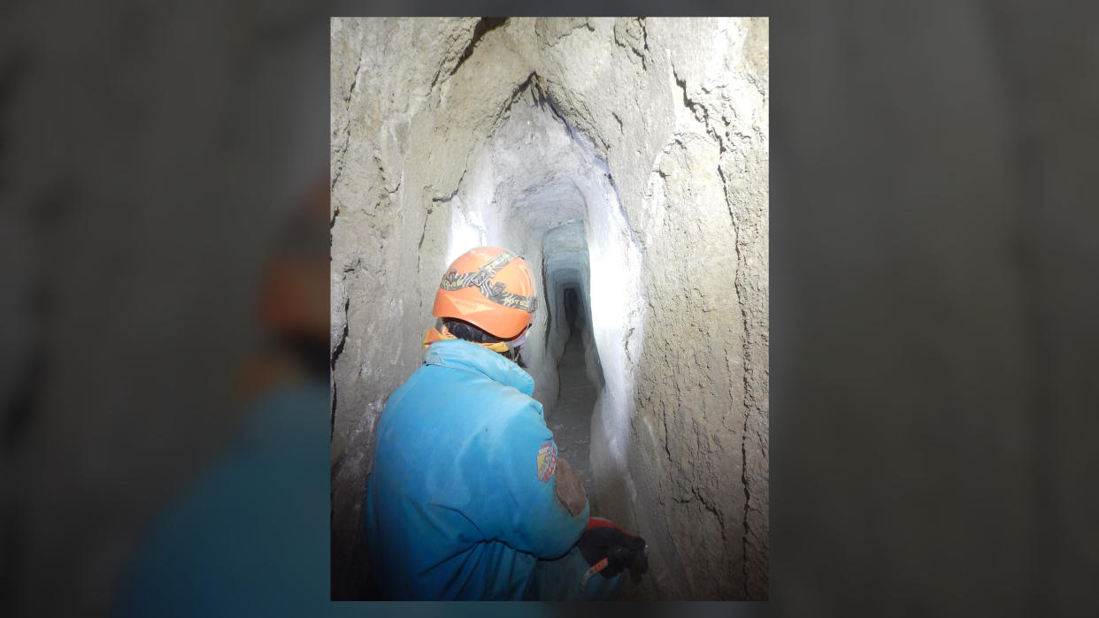  A man in a blue coat and orange helmet explored a narrow, dark cave-like aqueduct with his flashlight. 